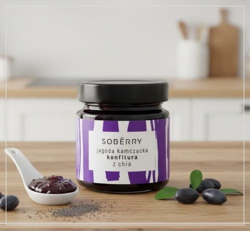 Szklany słoik konfitury z jagody kamczackiej z nasionami chia marki Soberry, ustawiony na jasnym dębowym blacie obok białej ceramicznej łyżeczki z konfiturą i świeżych owoców haskap